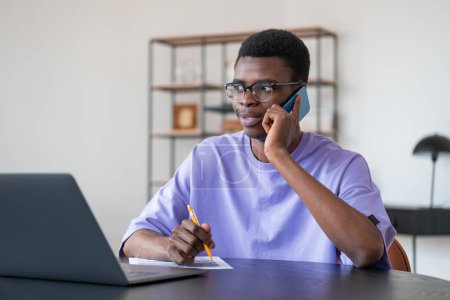 Téléchargez les photos : Un homme d'affaires africain au téléphone, ordinateur portable avec contrat sur le bureau. Salle de bureau avec étagère sur fond. Concept d'accord et de négociation - en image libre de droit