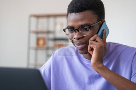 Téléchargez les photos : Un homme d'affaires africain souriant appelle au téléphone, regardant l'écran de l'ordinateur portable. Salle de bureau avec étagère sur fond flou. Concept de communication en ligne - en image libre de droit