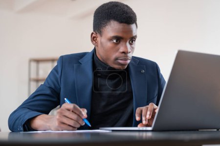 Photo for Black businessman looking at laptop screen and take note in paper, concentrated and pensive look. Concept of web courses and career development - Royalty Free Image