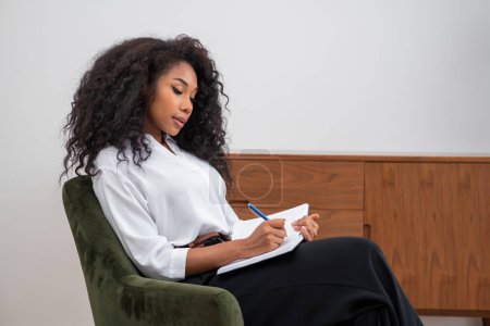 Konzentrierte schwarze Geschäftsfrau notiert im Notizbuch, im Sessel sitzend. Umfassendes Frauenprofil im minimalistischen Büroraum. Gründungskonzept und Geschäftsidee