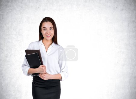 Foto de Mujer de negocios sonriente sosteniendo diario, mirando a la cámara con una sonrisa feliz, fondo de pared de hormigón gris. Concepto de aprendizaje y carrera. Copiar espacio - Imagen libre de derechos