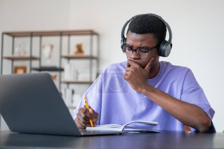 Foto de El estudiante afroamericano que usa ropa casual está sentado escuchando podcast a través de auriculares y computadoras portátiles y tomando notas en un cuaderno. Concepto de distante, en línea, aprendizaje remoto, educación empresarial - Imagen libre de derechos