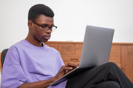 Téléchargez les photos : Un homme d'affaires afro-américain sérieux portant des vêtements décontractés travaille sur un ordinateur portable le tenant sur ses genoux. Concept d'enseignement à distance, en ligne, à distance, en entreprise - en image libre de droit