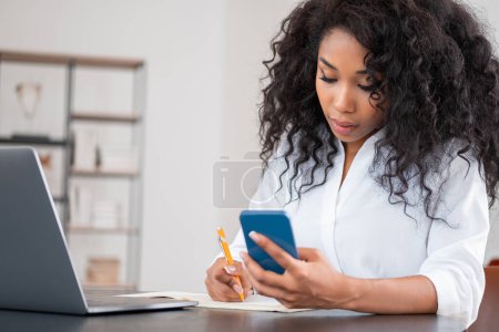 Photo for African businesswoman take note with mobile phone in hand, laptop and notebook on desk. Office room with shelf on background. Concept of distance work - Royalty Free Image