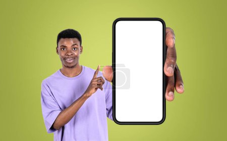 Foto de Joven hombre negro sonriendo, señalando con el dedo al teléfono inteligente, gran simulacro de pantalla de espacio de copia en el fondo verde. Concepto de red en línea y aplicación móvil - Imagen libre de derechos