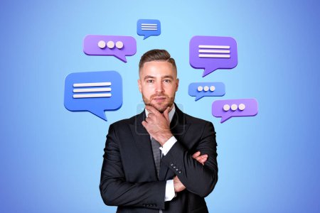 Photo for Portrait of pensive young bearded businessman with speech bubbles around him over blue background. Concept of communication and social media - Royalty Free Image