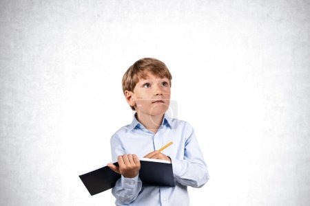 Téléchargez les photos : Portrait d'un petit garçon sérieux écrit dans un cahier noir avec un stylo sur un fond mural en béton. Concept d'éducation et d'étude - en image libre de droit