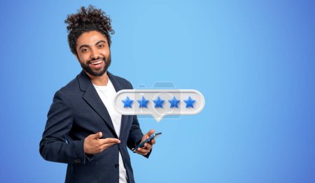 Photo for Happy arab businessman pointing at phone and five stars rating, empty copy space blue background. Good review from customer and positive feedback. Concept of rating and online service - Royalty Free Image