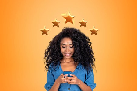 Foto de Mujer joven negro mensajes de texto en el teléfono en las manos, fondo naranja. Dar comentarios de cinco estrellas y compartir su experiencia positiva en línea. Concepto de servicio al cliente y calificación - Imagen libre de derechos