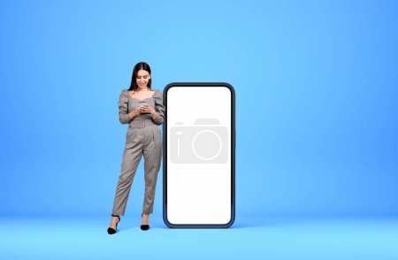 Téléchargez les photos : Attrayant femme d'affaires portant des vêtements formels est debout tenant smartphone près du mur bleu vide et un écran géant en arrière-plan. Concept de processus de travail, communication internet - en image libre de droit
