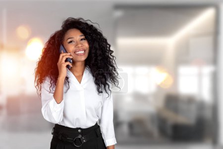 Photo for Attractive African American businesswoman wearing formal wear is standing talking on smartphone at office workplace in background. Concept of working process, important conference call - Royalty Free Image