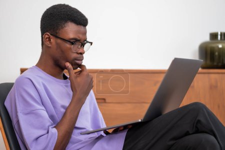Foto de Un hombre de negocios afroamericano serio que usa ropa casual está sentado trabajando en una computadora portátil sosteniéndola en su regazo y tocándose la barbilla. Concepto de distante, en línea, aprendizaje remoto, educación empresarial - Imagen libre de derechos