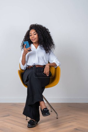 Photo for African businesswoman sitting in armchair, smiling and texting on the phone in hand. Concept of social media and communication - Royalty Free Image