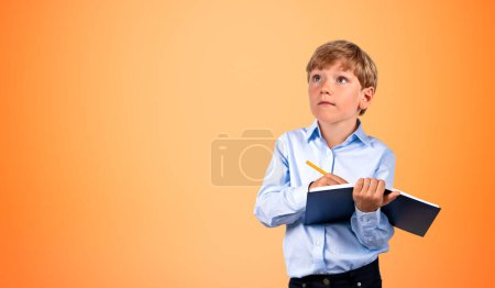 Foto de Retrato de un niño serio escribiendo en cuaderno negro con bolígrafo sobre fondo naranja. Concepto de educación y estudio. Prepárate. - Imagen libre de derechos