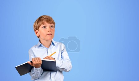 Téléchargez les photos : Portrait d'un petit garçon sérieux écrivant dans un cahier noir avec un stylo sur fond bleu. Concept d'éducation et d'étude. Maquette - en image libre de droit