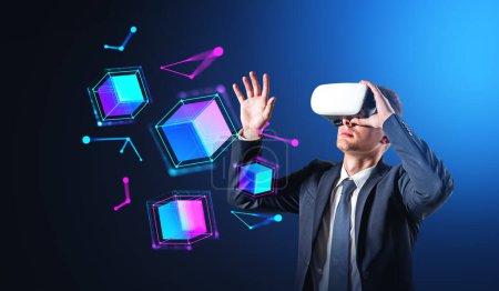 Téléchargez les photos : Portrait de jeune homme d'affaires en lunettes VR utilisant une interface métaverse sur fond bleu foncé. Concept de cyberespace et de haute technologie - en image libre de droit