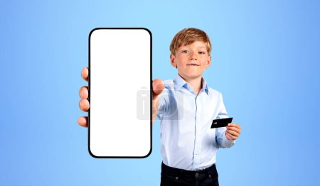 Téléchargez les photos : Enfant tenant smartphone avec écran vide maquette, tenant une carte de crédit à la main sur fond bleu. Concept de paiement, de transaction et d'achat en ligne - en image libre de droit
