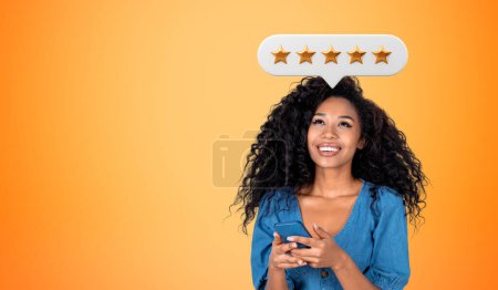Photo for Happy black woman with phone in hands, empty copy space orange background. Positive review from customer, giving five stars bubble feedback online. Concept of rating and best service - Royalty Free Image