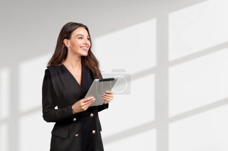 Photo for Attractive businesswoman wearing formal wear is standing holding tablet device near empty white wall in background. Concept of working process, mobile communication, time management - Royalty Free Image