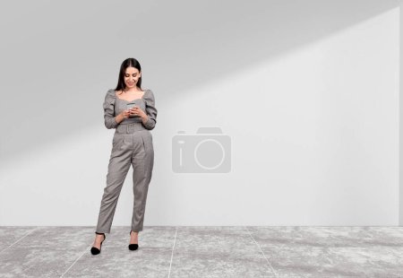 Foto de Atractiva mujer de negocios que usa ropa formal está de pie sosteniendo el teléfono inteligente cerca de la pared vacía en el fondo. Piso de hormigón. Concepto de proceso de trabajo, comunicación por Internet, gestión del tiempo - Imagen libre de derechos