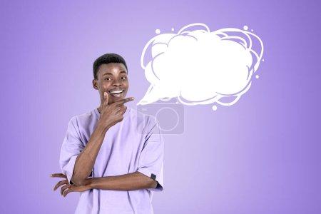 Téléchargez les photos : Jeune homme noir en t-shirt violet, souriant avec un regard inspiré, doigt pointant vers la bulle de pensée nuage blanc maquette sur fond violet. Concept du nouveau plan - en image libre de droit