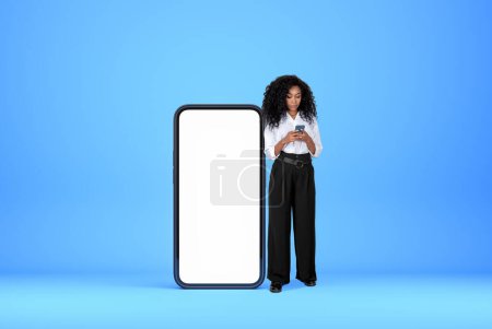 Téléchargez les photos : Femme d'affaires noire tapant dans le smartphone, pleine longueur près de grand écran de téléphone maquette, fond bleu clair. Concept de médias sociaux et de communication - en image libre de droit