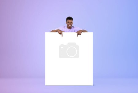 Photo for Black smiling man fingers pointing at blank square whiteboard. Mockup canvas on purple gradient background. Concept of presentation and recommend - Royalty Free Image