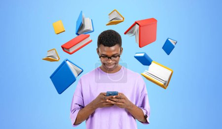 Foto de Sonriente hombre africano escribiendo en el teléfono vistiendo camiseta púrpura, flotando libros de colores sobre fondo azul. Concepto de biblioteca digital, educación, aplicación móvil y e-learning - Imagen libre de derechos