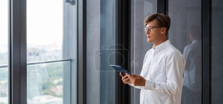 Foto de Hombre de negocios concentrado en gafas con la tableta en las manos, de pie cerca de la ventana panorámica con vista a la ciudad. Concepto de compras en línea, Internet y trabajo remoto - Imagen libre de derechos