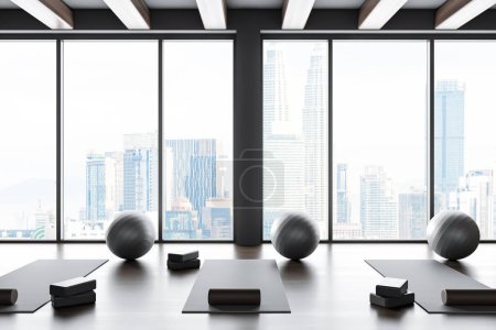 Téléchargez les photos : Intérieur salle de gym yoga sombre avec tapis et fitball avec bloc et rouleau, plancher de bois franc. Salle de classe de sport avec équipement et fenêtre panoramique sur Kuala Lumpur. rendu 3D - en image libre de droit