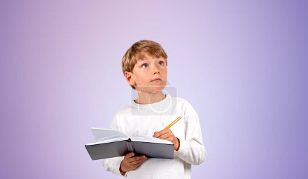 Téléchargez les photos : Enfant rêveur avec stylo prenant des notes dans un carnet, portrait souriant regardant vers le haut sur fond violet vide. Concept d'éducation, d'apprentissage et d'études en ligne - en image libre de droit