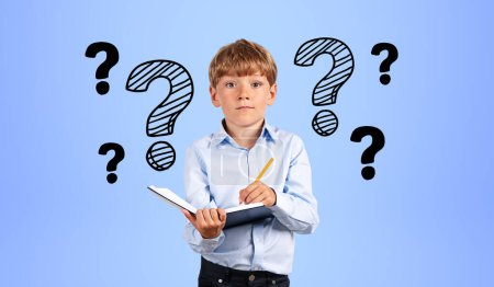 Photo for Portrait of serious little boy in blue shirt writing in notebook near blue wall with question marks drawn on it. Concept of curiosity and education - Royalty Free Image