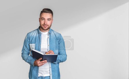 Téléchargez les photos : Jeune homme barbu prenant des notes dans un carnet, portrait regardant la caméra sur l'espace de copie fond d'ombre vide. Concept de gestion du calendrier, des idées et du temps - en image libre de droit