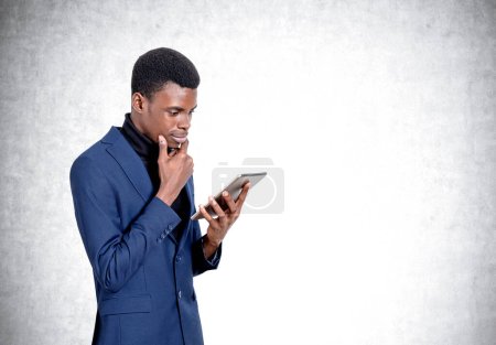 Foto de Hombre de negocios afroamericano guapo con traje formal está de pie mirando en el dispositivo de la tableta cerca de la pared de hormigón vacío en el fondo. Concepto de aparatos modernos, comunicación móvil, gestión del tiempo - Imagen libre de derechos