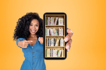 Téléchargez les photos : Jeune femme noire sourire doigt pointant vers grand smartphone avec bibliothèque numérique sur fond jaune. Concept d'apprentissage et d'éducation en ligne - en image libre de droit
