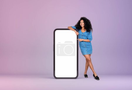Foto de Mujer joven negro sonriendo, de cuerpo entero, apuntando a un gran teléfono inteligente simulan pantalla en blanco sobre fondo púrpura. Concepto de redes sociales y comunicación - Imagen libre de derechos