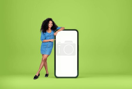 Téléchargez les photos : Jeune femme noire souriante, pleine longueur, pointant vers le grand smartphone maquette écran blanc sur fond vert. Concept de médias sociaux et de réseau - en image libre de droit