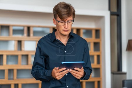 Téléchargez les photos : Jeune homme d'affaires sérieux en chemise bleue à l'aide d'un ordinateur tablette debout près du mur blanc et en verre. Concept de communication - en image libre de droit