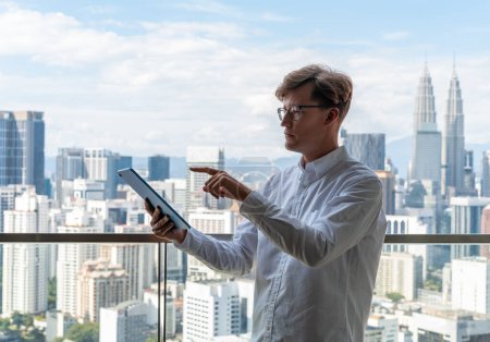 Foto de Retrato de un joven empresario europeo serio con camisa blanca usando una tableta de pie cerca de una ventana panorámica. Concepto de comunicación - Imagen libre de derechos