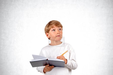 Téléchargez les photos : Enfant de l'école intelligente avec stylo prenant des notes dans un cahier, portrait intéressé regardant vers le haut sur fond de mur en béton gris vide. Concept de connaissance, d'éducation et de choix de carrière - en image libre de droit