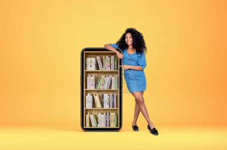 Téléchargez les photos : Jeune femme noire pleine longueur, souriante et pointant vers un grand smartphone avec des livres numériques sur fond jaune. Concept d'apprentissage en ligne - en image libre de droit
