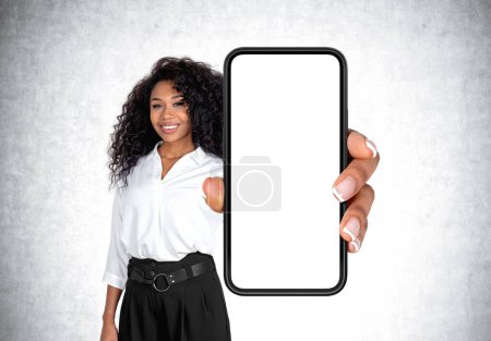 Téléchargez les photos : Femme d'affaires afro-américaine en tenue formelle tient smartphone avec de l'espace de copie debout devant un mur en béton en arrière-plan. Mockup screen, Présentation de l'application mobile et des médias sociaux - en image libre de droit