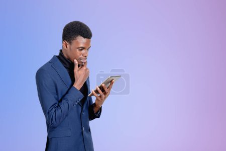 Téléchargez les photos : Beau homme d'affaires afro-américain portant un costume formel est debout en regardant l'appareil tablette près du mur violet vide en arrière-plan. Concept de gadgets modernes, communication mobile, gestion du temps - en image libre de droit
