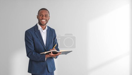 Foto de Retrato de un joven y alegre empresario afroamericano con un elegante traje que sostiene un cuaderno y un bolígrafo y está de pie cerca de una pared blanca. Concepto de planificación. Copiar espacio - Imagen libre de derechos