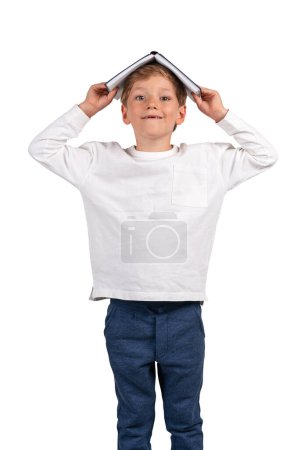 Téléchargez les photos : Écolier souriant et debout avec livre sur la tête, regardant la caméra, isolé sur fond blanc. Concept d'éducation, de carrière, d'idée et d'inspiration - en image libre de droit