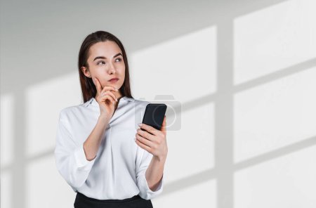 Photo for Pondering businesswoman wearing formal wear is standing touching chin holding smartphone near empty white wall in background. Concept of model, successful businessperson, thinking about business - Royalty Free Image