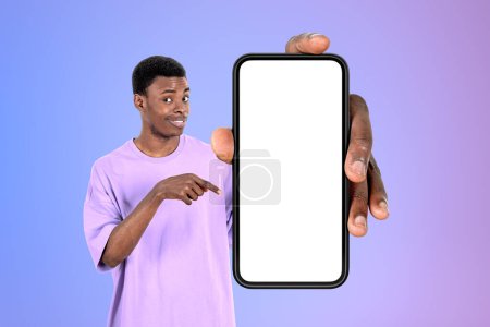Téléchargez les photos : Jeune homme noir souriant, doigt pointant vers le grand smartphone maquette écran d'espace de copie, fond dégradé violet. Concept de suivre et de souscrire - en image libre de droit