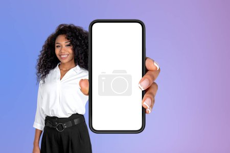 Téléchargez les photos : Femme d'affaires afro-américaine portant des vêtements formels, tenant smartphone avec écran de maquette, debout devant le mur dégradé violet en arrière-plan. Présentation de l'application mobile et des médias sociaux - en image libre de droit