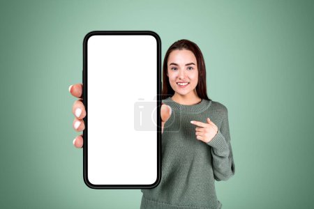 Photo for Young woman smiling, finger pointing at large smartphone mock up copy space screen, green background. Concept of new mobile app - Royalty Free Image