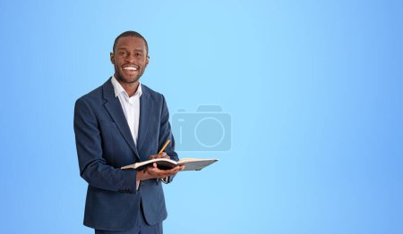 Photo for Portrait of cheerful young African American businessman in elegant suit holding notebook and pen and standing near blue wall. Concept of planning. Copy space - Royalty Free Image